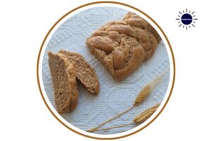 Wholegrain Spelt Vegan Challah On A Lace Cloth With Decorative Wheat On The Side Within A Decorative Circle With The Kosher Create Sun Logo On The Right Top.