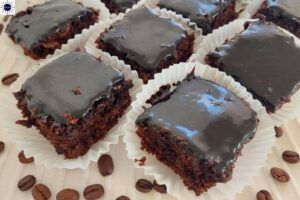 Wholegrain Almond Mocha Brownies With A Mocha Glaze On Top In Cupcake Holders Spread On A Pleated Cloth With Coffee Beans Scattered Around.