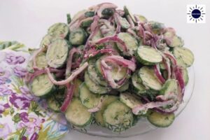 Mediterranean No-Mayo Cucumber Salad In Glass Serving Bowl With A Colorful Flowered Cloth Napkin On The Side And The Kosher Create Sun Logo In The Upper Right Corner.
