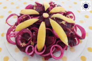 Mediterranean Lemony Beet Salad On A Glass Serving Plate Garnished With Onions Cut In Circles And A Large Petal Flower Carved From A Lemon Peel In The Center And With The Kosher Create Sun Logo On The Upper Right Side.