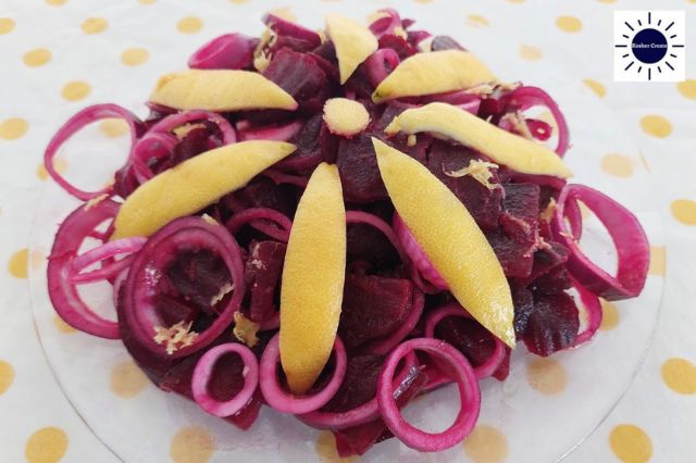 Mediterranean Lemony Beet Salad On A Glass Serving Plate Garnished With Onions Cut In Circles And A Large Petal Flower Carved From A Lemon Peel In The Center And With The Kosher Create Sun Logo On The Upper Right Side.