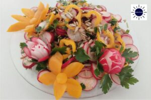 Mediterranean Crunchy Radish Salad On A Glass Serving Plate Adorned With A Carved Radish Rose And Two Flowers Carved From Peppers With The Kosher Create Sun Logo On The Upper Right Side.