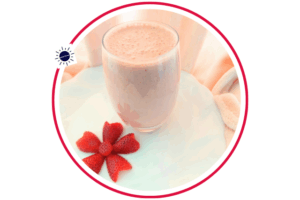 Strawberry Peanut Butter Yogurt Smoothie In A Glass On A Ruffled Rim Plate Adorned With A Flower Carved Out Of A Strawberry With A Gathered Textured Cloth In The Background That’s Suited For Breakfast Within A Decorative Circle Adorned With The Kosher Create Sun Logo.