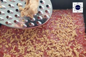 Grating The Cold Dough With A Grater Over The Strawberry Filling In The Baking Pan With The Kosher Create Sun Logo In The Upper Right Corner.
