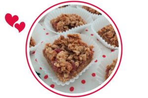 Wholegrain Strawberry Peanut Butter Bars In Cup Cake Holders Spread Out On A Doted Cloth That's Suited For Dessert In A Decorative Circle Adorned With Two Graphic Hearts On The Side.