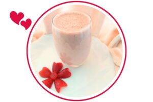 Strawberry Peanut Butter Yogurt Smoothie In A Glass On A Ruffled Rim Plate Adorned With A Flower Carved Out Of A Strawberry With A Gathered Textured Cloth In The Background That's Suited For Dessert In A Decorative Circle Adorned With Two Graphic Hearts On The Side.