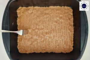 Peanut Butter Dough On Bottom Of Baking Pan With Holes Perforated With A Fork With The Kosher Create Sun Logo In The Upper Left Corner.