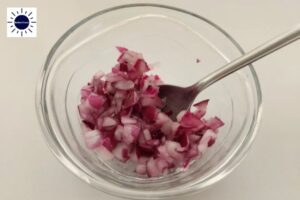 Cubed Red Onion Mixed With Salt With A Spoon In A Glass Bowl With The Kosher Create Sun Logo In The Upper Left Corner.