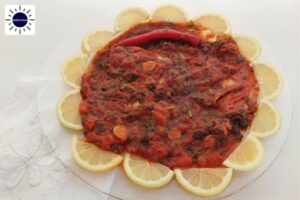 Baked Mediterranean Tomatoey Spicy Fish On A Round Glass Serving Plate Adorned With Fresh Lemon Wedges Around That Creates A Flower With a Decorative Cloth Napkin On The Bottom Left Side And The Kosher Create Sun Logo In The Upper Left Corner.