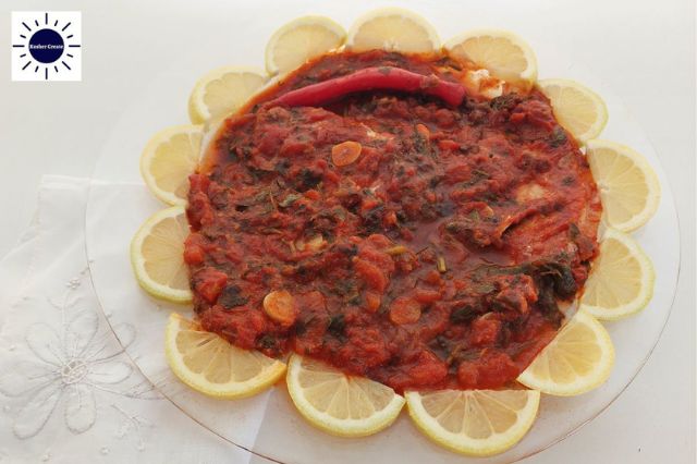 Baked Mediterranean Tomatoey Spicy Fish On A Round Glass Serving Plate Adorned With Fresh Lemon Wedges Around That Creates A Flower With a Decorative Cloth Napkin On The Bottom Left Side And The Kosher Create Sun Logo In The Upper Left Corner.