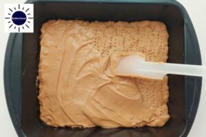 Peanut Butter Spread With A Spatula On The Baked Base Dough In A Baking Pan With The Kosher Create Sun Logo In The Upper Left Side.