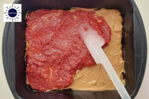 Strawberry Filling Spread With A Spatula On The Peanut Butter Layer In The Baking Pan With The Kosher Create Sun Logo In The Upper Left Corner.