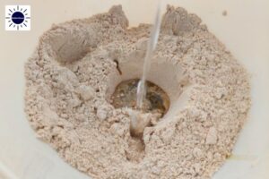 Closeup Of Water Pouring Into The Well Created In The Flours With The Kosher Create Sun Logo In The Upper Left Corner.