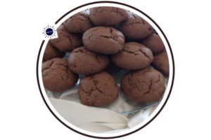Wholegrain Cocoa Maple Mocha Cookies On A Glass Serving Plate On Top Of Bunched Silky Material With a Decorative Silky Large Bow In Front That's Suited For Breakfast In A Decorative Circle Adorned With The Kosher Create Sun Logo.