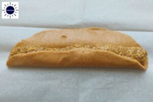 A Roll Of Filling Placed In The Middle Of The Oval Shaped Dough Lengthwise With The Front Part Of The Dough Folded To The Middle On A Baking Sheet With The Kosher Create Sun Logo In The Upper Left Corner.