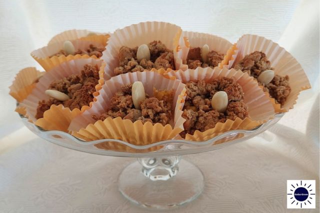Wholegrain Almond Fresh Apricot Bars Within A Decorative Cupcake Form With A Blanched Almond In The Center Arranged In A Circular Fashion On A Glass Cake Stand With The Kosher Create Sun Logo In The Right Lower Corner.