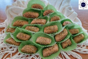 Mediterranean Wholegrain Pistachio Cookies Arranged In A Pyramid With Each Cookie In A Decorative Cupcake Holder With Curled Tassels In The Front And A Bunched Textured Cloth In The Back With The Kosher Create Sun Logo In The Upper Right Side.