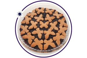 A Closeup Of A Wholegrain Lemony Blueberry Pie Decorated With Blueberry Cookies On Top In A Round Glass Pie Dish On Top Of A Lace Cloth With Blueberry Ornaments That's Suited For Breakfast In A Decorative Circle Adorned With The Kosher Create Sun Logo.