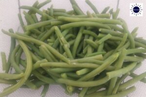 A Closeup Of Cooked Green Beans Drained In A Sieve With The Kosher Create Sun Logo In The Upper Right Corner.