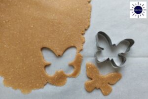 A Cookie Form And A Cookie Cut From Dough On A Baking Sheet With The Kosher Create Sun Logo In The Upper Right Side.