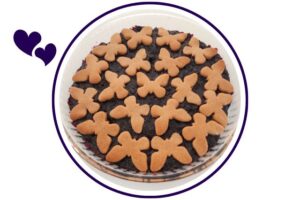 A Closeup Of A Wholegrain Lemony Blueberry Pie Decorated With Blueberry Cookies On Top In A Round Glass Pie Dish On Top Of A Lace Cloth With Blueberry Ornaments That's Suited For Dessert In A Decorative Circle Adorned With Two Graphic Hearts On The Side.
