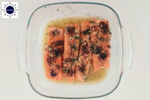 Pesto Spread Over The Salmon Slices Within A Square Glass Baking Dish With The Kosher Create Sun Logo In The Upper Left Corner.