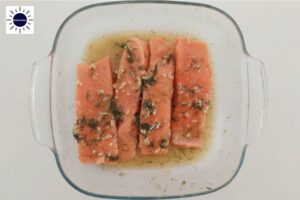 The Salmon Slices Marinated In A Deep Glass Square Dish With The Kosher Create Sun Logo In The Upper Left Corner.