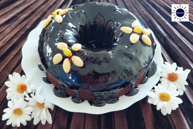 Wholegrain Dairy-Free Glazed Cocoa Cake Adorned With Three Flowers Made Out Of Blanched Almonds On A Glass Serving Plate With Two Groups Of Cloth Daisies On The Side On Top Of A Pleated Textured Cloth With The Kosher Create Sun Logo In The Upper Right Corner.