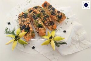Mediterranean Baked Lemony Honey Salmon On A Round Glass Serving Dish Adorned With Diced Fresh Dill And With Nigella seeds And With Two Decorative Flowers Carved From Lemon Peels In The Front On Top Of A Decorative Cloth With The Kosher Create Sun Logo In The Upper Right Corner.