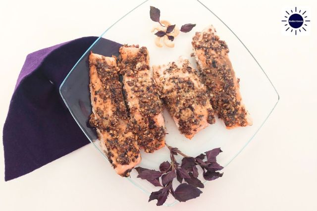 Mediterranean Baked Cashew Pesto Salmon On A Diamond Shaped Glass Serving Plate Adorned With Red Basil Leaves And A Flower Made Out Of Cashews With A Decorative Cloth On The Side Shaped Like A Basil Leaf And With The Kosher Create Sun Logo In The Upper Right Side.