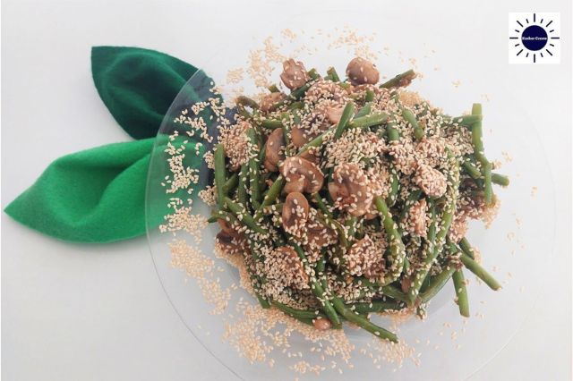 Mediterranean Savory Mushroom Green Beans On A Glass Serving Plate Adorned With Roasted Sesame Seeds And With Two Cloth Leaves On The Upper Left Side And With The Kosher Create Sun Logo In The Upper Right Corner.