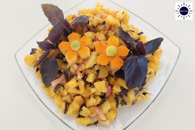 Mediterranean Baked Purple Potato Salad In A Diamond Shaped Glass Dish Adorned With Two Flowers Made Ot Of Potatoes Sprinkled With Turmeric And Basil Leaves On Their Sides And With The Kosher Create Sun Logo In The Upper Right Corner.