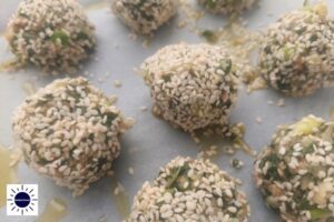 A Closeup Of Sesame Coated Chard Balls Placed On A Baking Sheet With The Kosher Create Sun Logo In The Lower Left Corner.