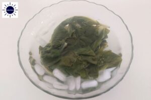 A Closeup Of The Cooked Chard Leaves Placed Into A Glass Bowl With Ice Water With The Kosher Create Sun Logo In The Upper Left Corner.
