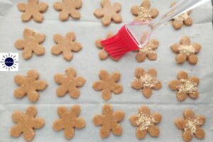 Cookie Forms Spread Out On A Baking Sheet Brushed With The Honey Water Mixture And Sprinkled With Chopped Cashews With The Kosher Create Sun Logo On The Upper Left Side.