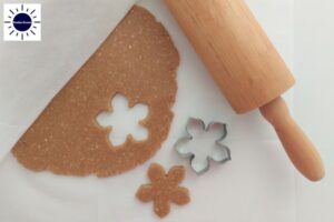 A Flower Cookie Shape Cut From The Dough With The Cookie Cutter And Rolling Pin On The Side And With The Kosher Create Sun Logo In The Upper Left Side.