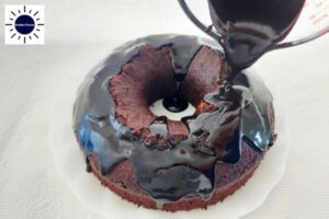 Pouring Glaze Onto The Cake That's On A Serving Plate With The Kosher Create Sun Logo In The Upper Left Corner.