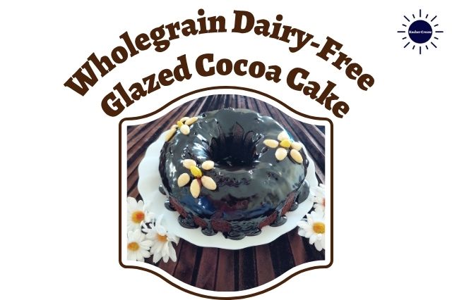 Wholegrain Dairy-Free Glazed Cocoa Cake Adorned With Three Flowers Made Out Of Blanched Almonds On A Glass Serving Plate With Two Groups Of Cloth Daisies On The Side On Top Of A Pleated Textured Cloth  Within A Decorative Frame With The Kosher Create Sun Logo In The Upper Right Corner.