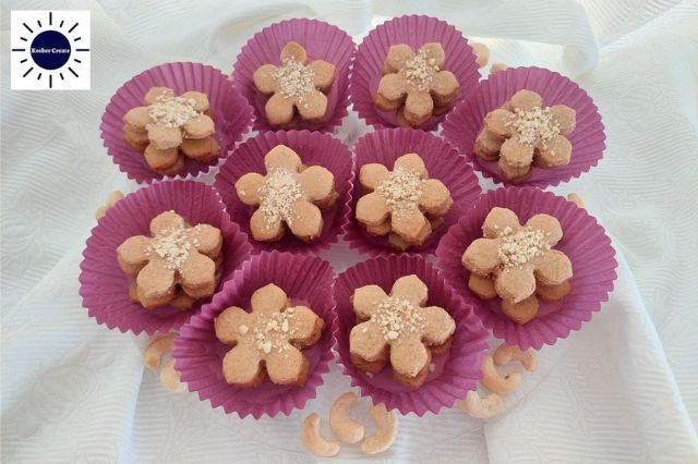 Wholegrain Lemony Cashew Cookies Stacked In Colorful Cupcake Holders Arranged On A Serving Plate Like A Flower With Assorted Cashew Nuts Around With The Kosher Create Sun Logo In The Top Left Corner.