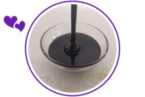 Tahini Cocoa Glaze Pouring Into A Glass Bowl On A Decorative Cloth With Sesame Seeds Scattered Around That's Suited For Dessert In A Decorative Circle Adorned With Two Graphic Hearts On The Side.