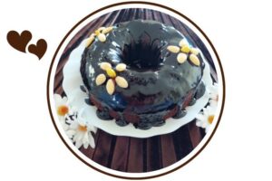 Wholegrain Dairy-Free Glazed Cocoa Cake Adorned With Three Flowers Made Out Of Blanched Almonds On A Glass Serving Plate With Two Groups Of Cloth Daisies On The Side On Top Of A Pleated Textured Cloth That's Suited For Dessert In A Decorative Circle Adorned With Two Graphic Hearts On The Side.   