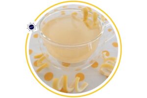 Lemoney Honey Ginger Drink In A Glass Round Mug Adorned With Fresh Lemon Swirls On The Mug And On The Polka Dot Cloth Beneath It Within A Decorative Circle Adorned With The Kosher Create Sun Logo.