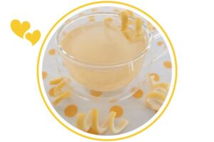 Lemoney Honey Ginger Drink In A Glass Round Mug Adorned With Fresh Lemon Swirls On The Mug And On The Polka Dot Cloth Beneath It That's Suited For Dessert In A Decorative Circle Adorned With Two Graphic Hearts On The Side.